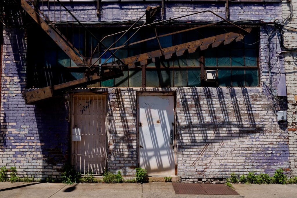 An urban building wall with faded bricks and peeling paint, rusting metal doors and iron bars, and a rusting fire escape casting shadows on the wall reflecting the passing time of day.