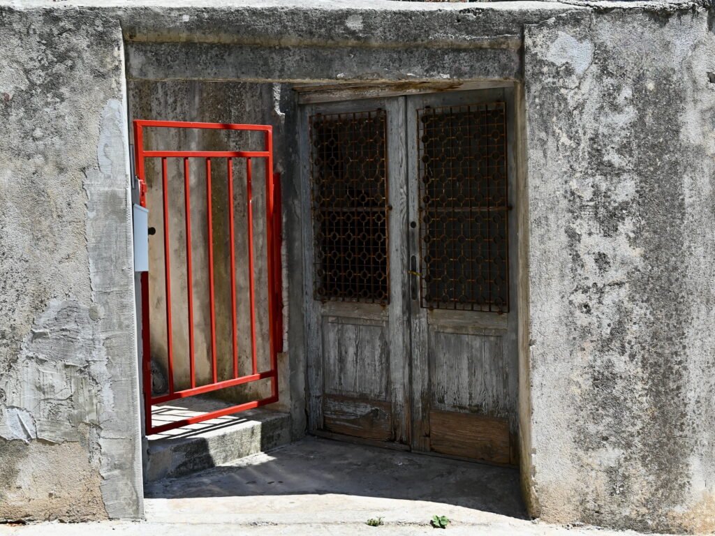 A building entrance in Split, Croatia, with cement and peeling plaster walls, a red metal gate, and old and weathered wooden double doors with rusty decorative metal grating.