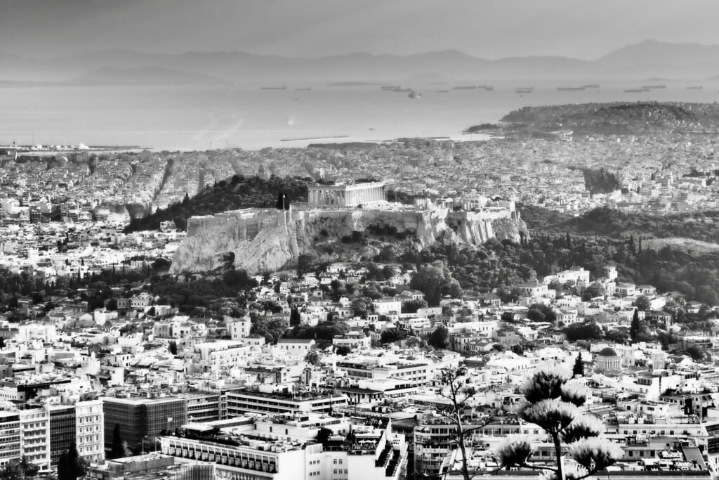 Black and white cityscape image of Athens with the Acropolis in the distance and ships anchored in the  Saronic Gulf in the background.