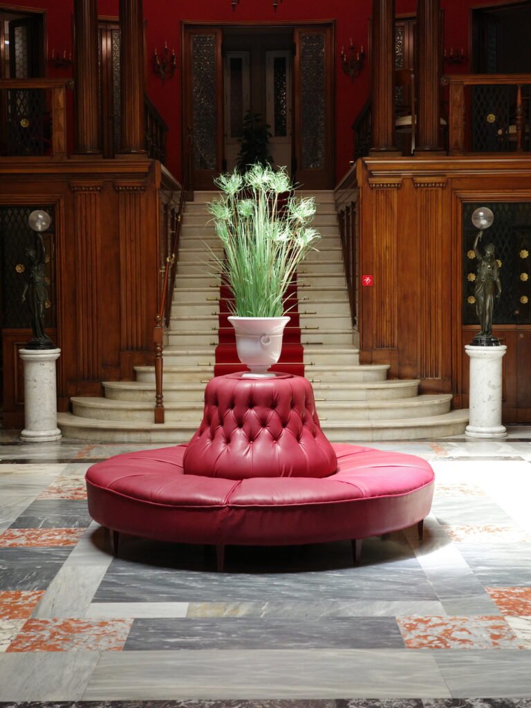 Elegant old world european hotel interior lobby with a round red leather couch and large vase in the center with a green plant. Ornate wood paneling and a white marble staircase with a red carpet running down the middle are in the background.