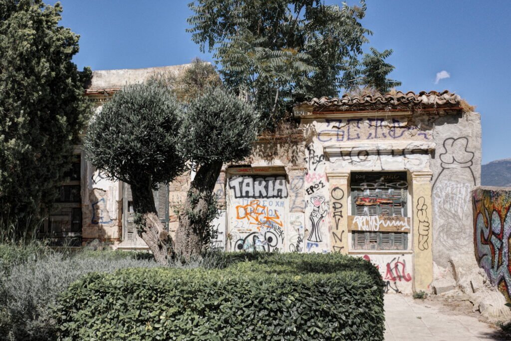 An old rustic home with weathered and crumbling walls covered with fading graffiti in Anafirotika, partially obscured by bushes and an olive tree, and expression of wabi-sabi.