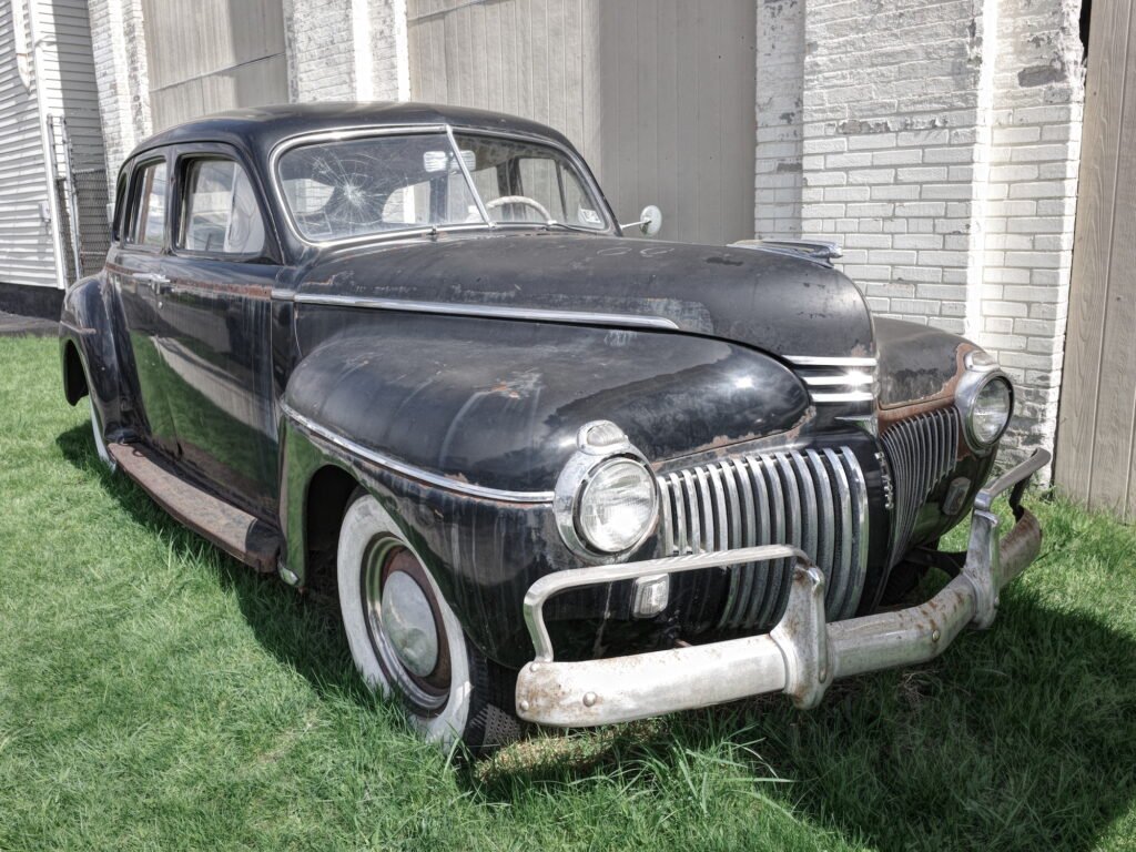 A vintage American car, 1940's era, with fading black paint, a cracked windshield, whitewall tires, and tarnished chrome bumpers and rusty patches parked on grass next to an old white brick wall.