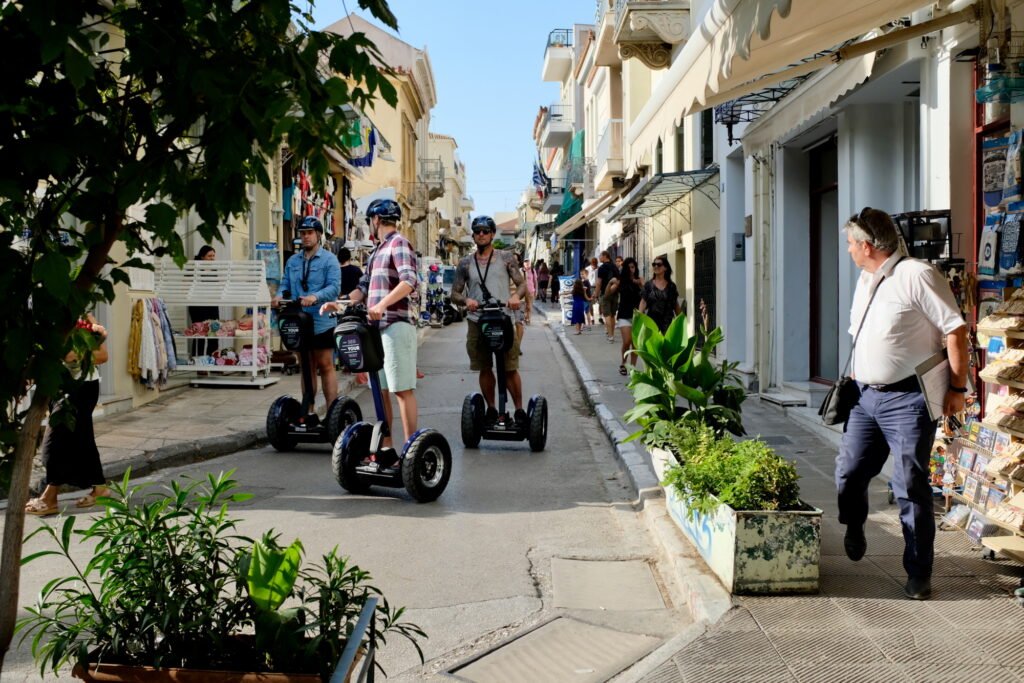 A busy alley in the Plaka in Athens, Greece.  Pedestrians fill the sidewalks while three helmeted riders on Segways ride down the center of the alley.