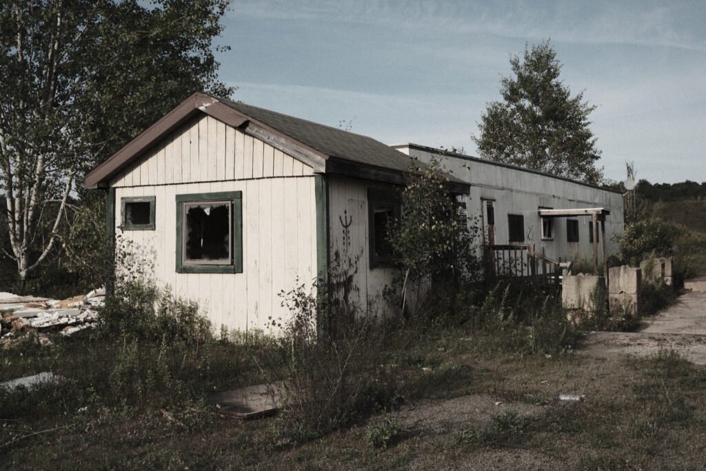 An old dilapidated and weathered building with broken windows and surrounded by weeds in a rural setting.