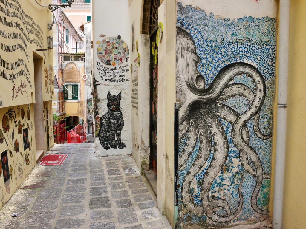 An old rustic narrow alley in Salerno, Italy, with artistic graffiti paintings on the wall with a large octopus and a black cat in the background.