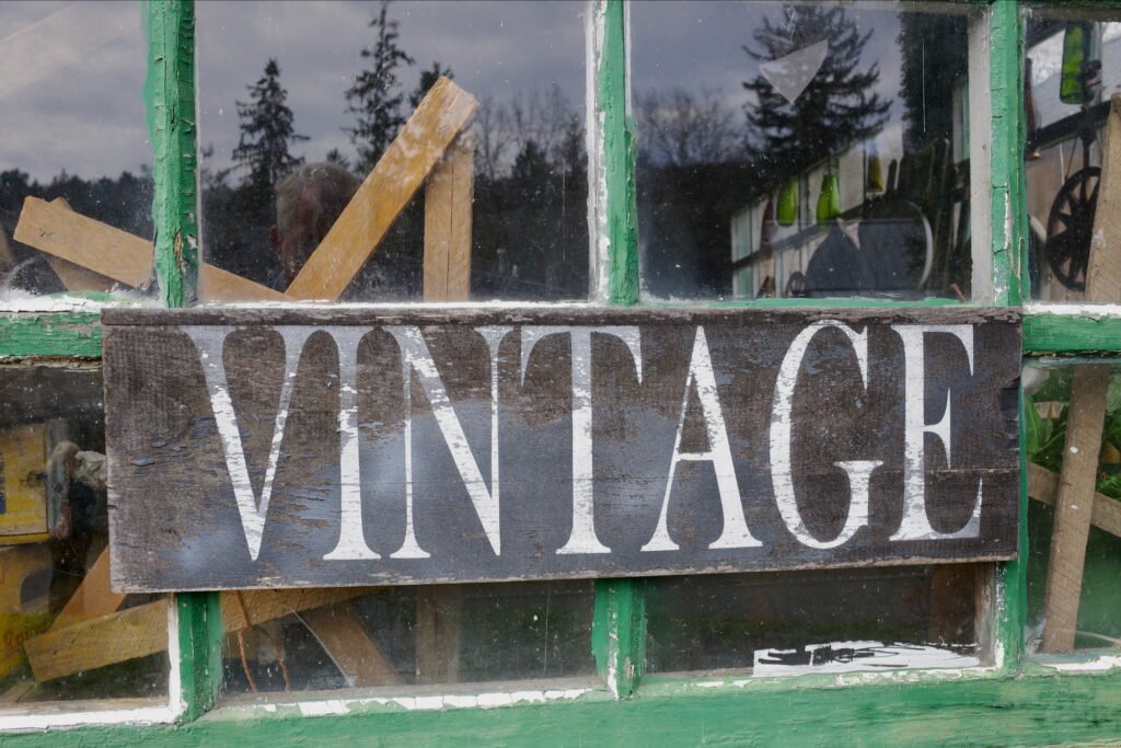 Old and rustic square pained window with peeling hunter green paint and a faded brown wood sign with the word vintage painted in fading white. Behind the windows in the background old wood and glass items can be seen.