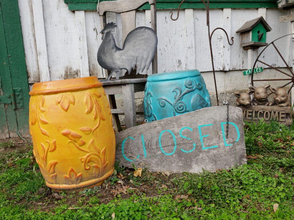Rural rustic image of vintage metal rooster, birdhouse, welcome sign with three pigs, one yellow and one blue old ornate barrels, and a hand-painted closed sign on weathered wood, with a weathered green and white wood wall with peeling paint in the background.