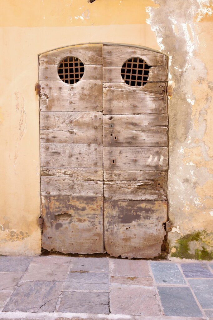 An old weathered wall with fading earth-toned colors and peeling plaster, with a tall narrow wood door with discolored wood planks and two small round windows at the top with rusty metal grates. At the foot of the door is a cracked and faded red and gray brick sidewalk.
