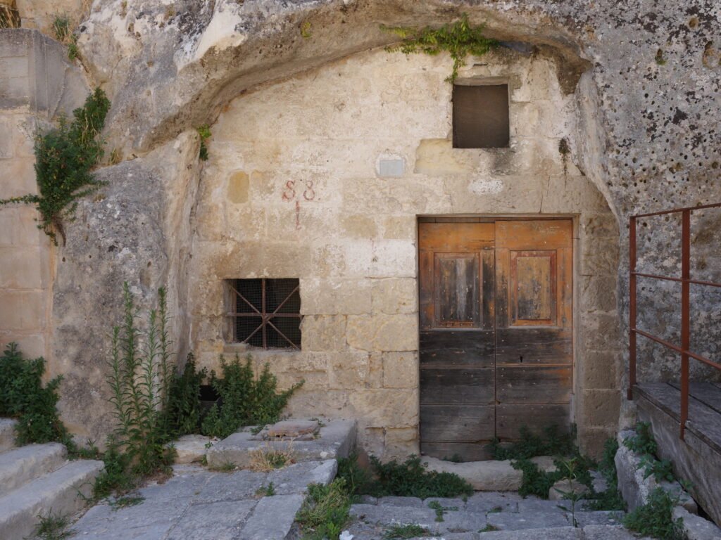 The entrance to a cave dwelling with a weathered wood door and a window with a rusty metal grate set directly in the ancient stone face of the mountain in the Stone City of Matera. Weeds grow out of the cracks in the stone walls and walkway in front of the door. The appearance is very rustic and a showcase of wabi-sabi aesthetics.