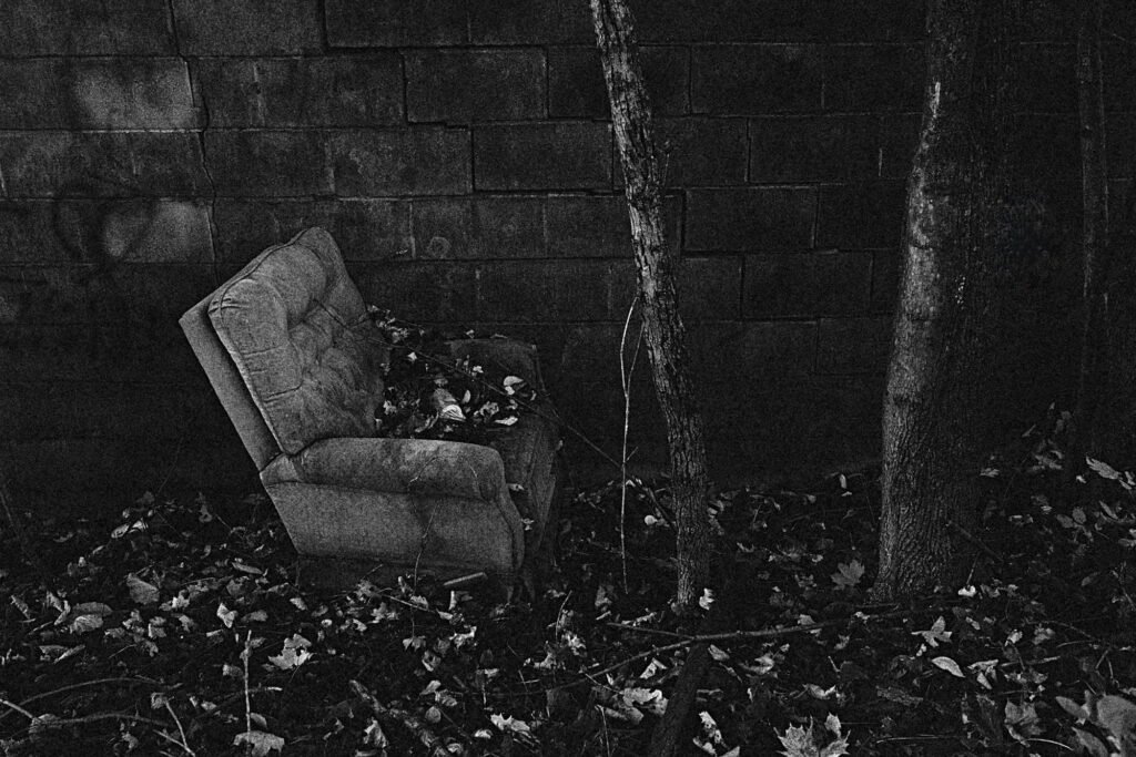 Gritty black and white image of urban or rural decay with an old easy chair sitting on ground covered with dried fallen leaves, in front of a textured cement block wall and next to two skinny bare tree trunks. Wabi-sabi grit and texture.
