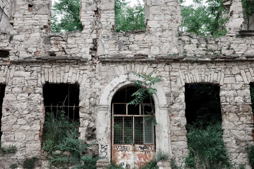 The facade of a crumbling war damaged stone building in Bosnia-Herzegovina with bushes and ferns growing through windowless and doorless openings as nature reclaims the space. A rusty and graffiti covered metal door is in the central arched opening to the building remains.
