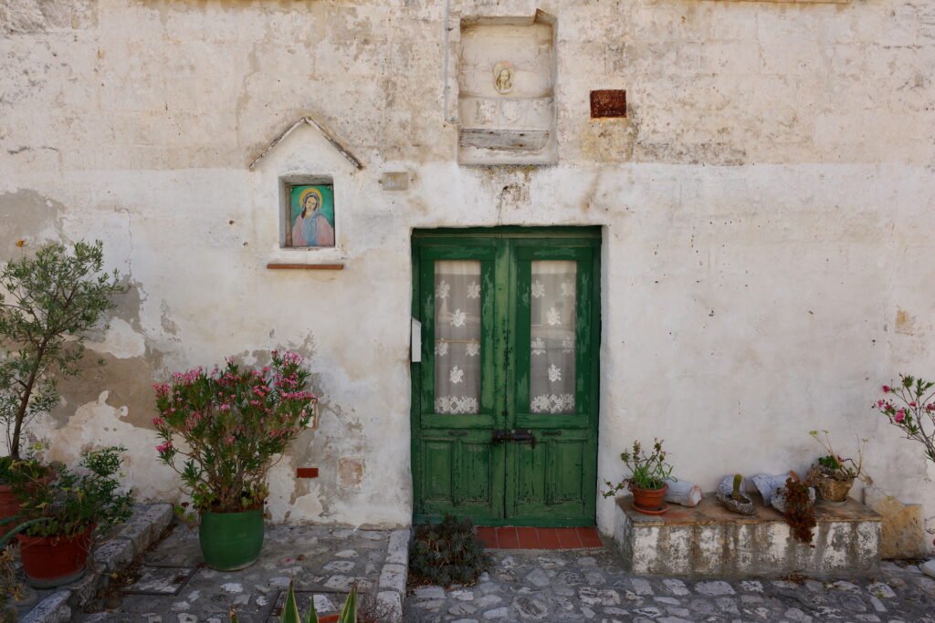 Weathered green double door entrance to a rustic home in the Stone City of Matera, Italy. The door is in an old and weathered wall with uneven stone steps and potted plants and flowers outside the door.