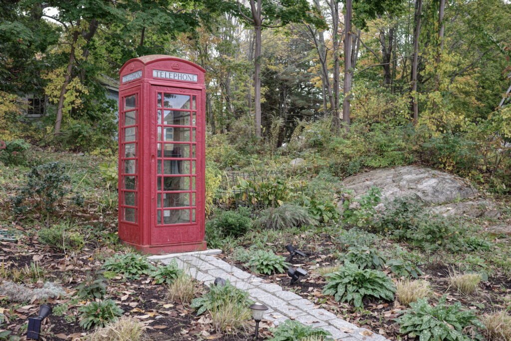 A classic old red telephone booth sits as a rural garden landscaping feature, surrounded by plants and trees, with a stone path leading to the telephone booth.