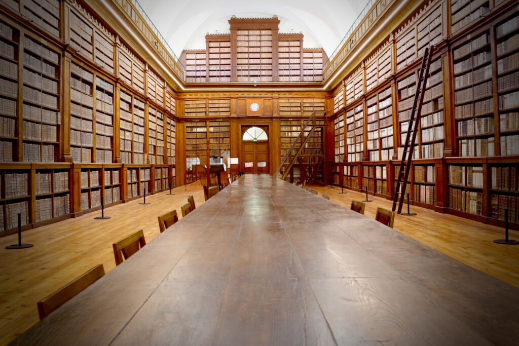 View inside an old and large library room looking down the length of a long wooden table with a door and clock behind it. Floor to ceiling book shelves and are also made of wood. Two ladders lean against the book stacks to reach the highest shelves. The room has a formal and old world look to it.