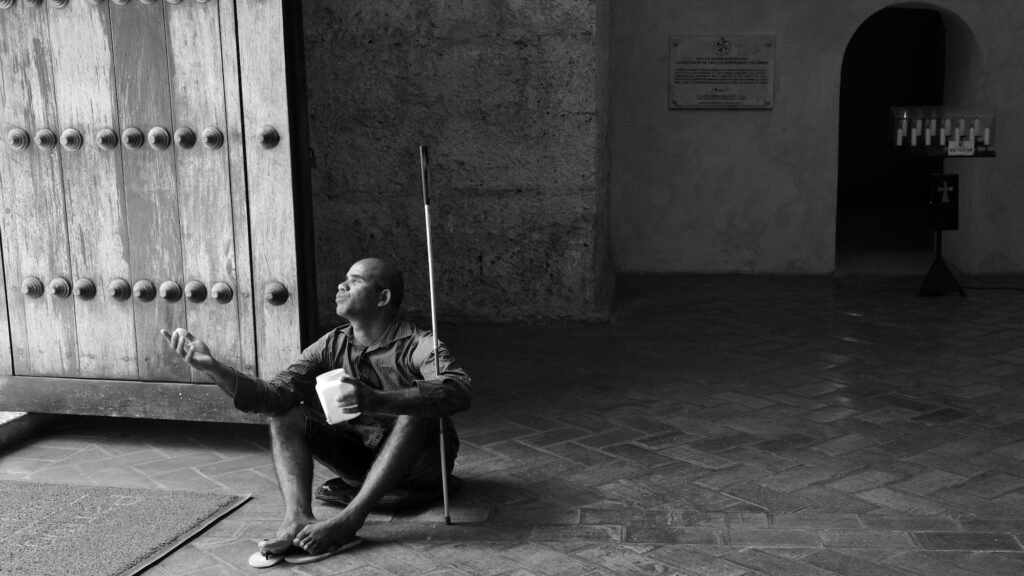 Black and white image of a blind panhandler with a cane sitting on a tile floor just inside a church entrance with light shining on him and the door as he holds a collection container in one hand and is reaching out with the other.