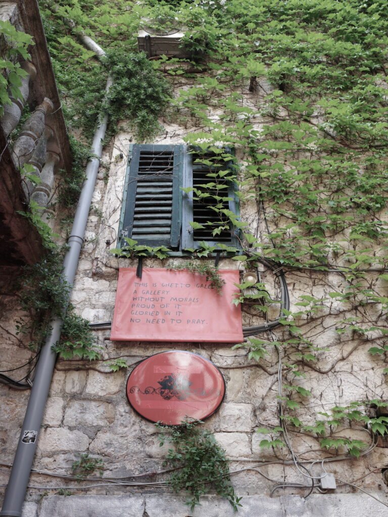Vines grow over an old earth-toned stone wall with a dilapidated and faded green shutter. Below the shutter is a red scroll with hand printed words that say This is Ghetto Gallery A Gallery Without Morals Proud of it Gloried in it No Need to Pray.