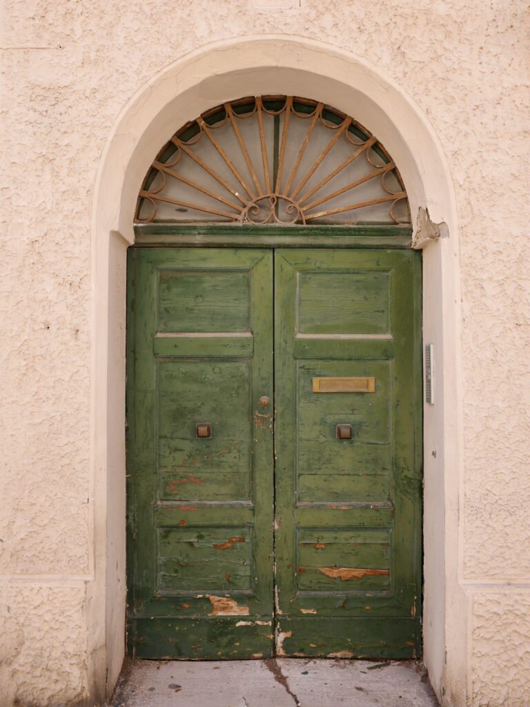 Green wooden door with weathered and peeling paint recessed in a weathered and stained brown stucco wall, with a decorative metal arch above the door.. Wabi-sabi aesthetics.