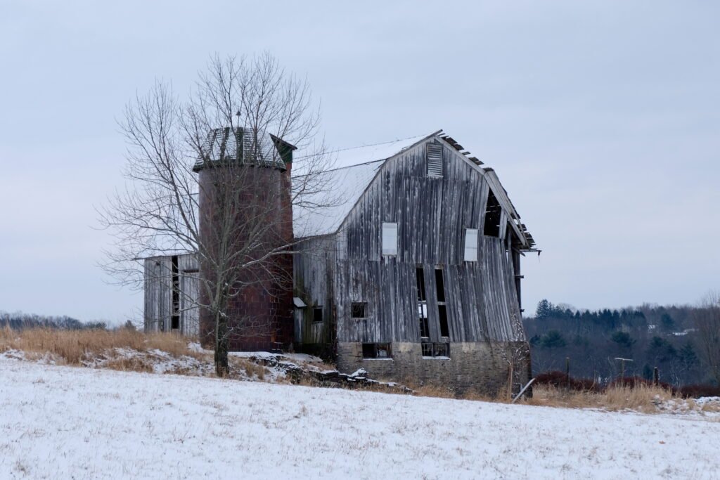 A silo and an old abandoned wooden barn with missing wood sections and  wall on the verge of collapse in a tranquil rural winter scene with a gray-blue sky, and light snow covering the ground.