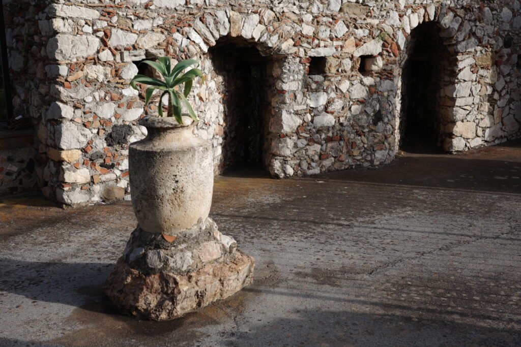 An ancient stone wall and outdoor vase with mixed natural stone and pottery pieces, with two open arched entryways in the wall. A green succulent plant grows in the vase.